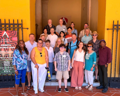 A colorful picture of a group of people in front of a yellow walled building with a sign beside them that reads