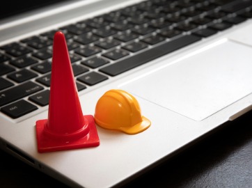Image of a the edge of a silver laptop with a small yellow construction hat and red traffic cone on top.