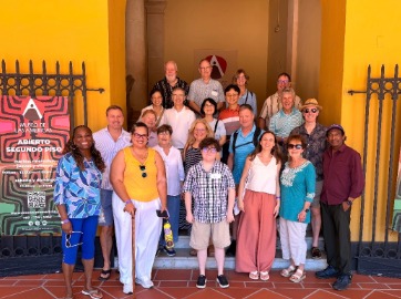 A colorful picture of a large group of individuals in front of a yellow walled building with a sign beside them that reads "Abierto Segundo Piso"
