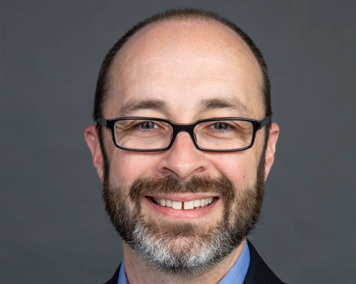An headshot of a man in a suit with a beard and glasses.
