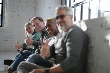 Four persons sit in a line looking at the camera with their thumbs up